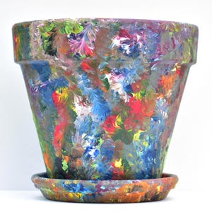 May include: A colorful, hand-painted ceramic flower pot with a matching saucer. The pot is painted with a vibrant mix of colors including red, blue, green, and yellow. The pot is approximately 8 inches tall and 9 inches wide.