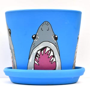 May include: A bright blue plant pot with a matching saucer, featuring a hand-painted illustration of a grey shark with a wide-open mouth revealing sharp white teeth and a vibrant pink interior, repeated around the exterior surface of the pot.