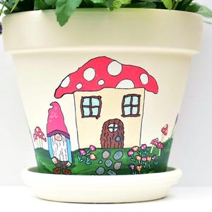 May include: Cream-colored ceramic flower pot with a whimsical hand-painted design. The design features a mushroom-shaped house with a red cap and white spots, a gnome, and colorful flowers. The pot sits on a matching saucer.