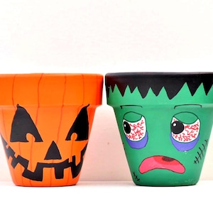May include: Two terracotta plant pots painted with Halloween themes. The left pot is orange with a black jack-o'-lantern face. The right pot is green with a black jagged hairline, bloodshot eyes, a pink mouth, and black stitches on the side.