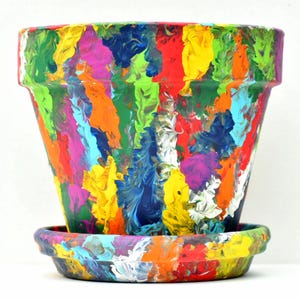 May include: A tapered terracotta plant pot and matching saucer featuring a vibrant, multi-colored abstract paint pour design. The surface displays thick, textured streaks of red, yellow, blue, green, orange, and purple acrylic paint.