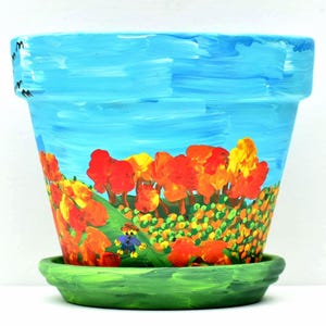 May include: A hand-painted terracotta plant pot with a matching saucer, featuring a vibrant autumn landscape with orange and yellow trees against a bright blue sky, and a small figure standing in a green field with tiny black birds in the corner.