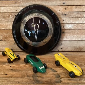 May include: A circular black and chrome V8 emblem featuring a stylized V8 logo in the center, positioned behind three wooden toy race cars. The cars include a yellow car with a green stripe, a green car with the number 88, and a yellow car with a checkered pattern.