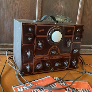 May include: A vintage brown cathode ray oscillograph featuring a central circular screen, multiple control knobs, and a top handle. The front panel displays the text "CATHODE RAY OSCILLOGRAPH" and "EXTERNAL OSCILLATOR" alongside two orange magazines.