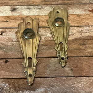 May include: Two matching brass door plates with a fan-shaped top and a vertical, tapered body. Each plate features a circular opening at the top for a knob and a keyhole opening below, with small screw holes at the top and bottom for mounting.