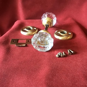 May include: A vintage door knob set with clear faceted glass knobs, brass hardware, and mounting screws. The set includes two knobs, two backplates, and a strike plate. The hardware is displayed on a red fabric.