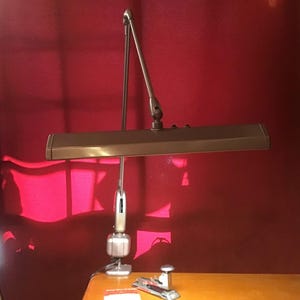 May include: A bronze-colored adjustable desk lamp with a rectangular light fixture and a silver-colored base. A silver stapler and a small notepad are on the wooden desk. The background is a solid red wall.