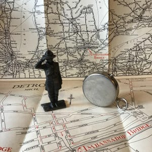 May include: A vintage map with a metal compass and a small, dark-colored figurine of a person saluting. The map shows the Detroit area and the Ambassador Bridge. The compass is silver-colored.