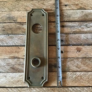May include: Antique brass door plate with two keyholes and decorative edges. The plate is approximately 6 inches tall.