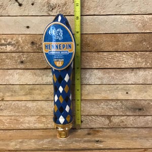 May include: A blue and gold beer tap handle with a diamond pattern. The handle features a circular label with the text "HENNEPIN FARMHOUSE SAISON BREWERY OMBRE  SCHLITZ  ALE".