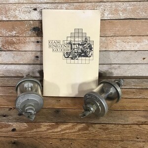 May include: An antique Gas Engine Guide book with a black and white illustration of a vintage vehicle. Two clear glass and metal fuel filters are in the foreground, with visible text. The items are on a wooden surface.