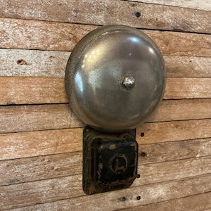 May include: A wall-mounted electric bell featuring a large, circular, metallic dome with a weathered finish and a central silver-toned nut. Below the dome is a rectangular, black-painted metal housing with visible signs of wear and paint loss.