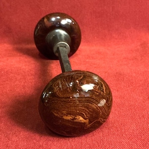 May include: A pair of round door knobs featuring a polished, dark brown marbled finish with swirling light brown patterns. The two knobs are connected by a central square metal spindle, showcasing a smooth, glossy surface texture throughout.