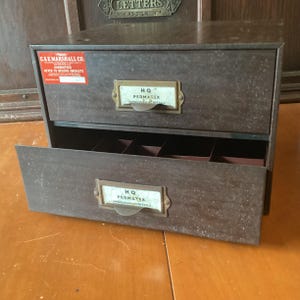 May include: Vintage metal storage cabinet with two drawers. The cabinet is a dark brown color with a distressed finish. Each drawer has a label holder with the text "HQ PERMATEX UNBREAKABLE CRYSTALS". A red sticker reads "C&E MARSHALL CO. GUARANTEED NEVER TO BECOME OBSOLETE".