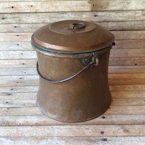 May include: A vintage copper bucket with a lid and a handle. The bucket is tarnished and has a rustic look.