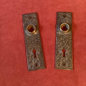 May include: Two rectangular metal door plates featuring an intricate embossed scrollwork pattern. Each plate includes a circular opening for a door handle, a keyhole cutout, and two small screw holes at the top and bottom for mounting purposes.