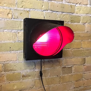 May include: A black, square traffic light with a single red lens illuminated. The light is mounted on a yellow brick wall. A black power cord extends from the bottom of the light.