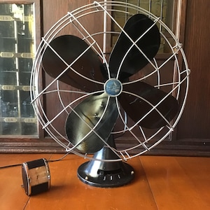 May include: Vintage black and chrome electric fan with a protective wire cage. The fan has four black blades and a round base. A separate cylindrical component is connected by a black cord. The fan is a classic home appliance.