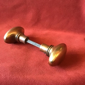 May include: A pair of oval-shaped brass door knobs connected by a central square metal spindle. The knobs feature a smooth, polished metallic finish with a warm golden hue, and each knob is attached to a circular base plate with a grooved edge.