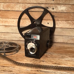 May include: A vintage black Kodak Fun Saver camera with a film reel and a film strip. The camera has a silver lens and a dial with the words "Fun Saver". The background is a wooden surface.