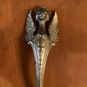May include: Close-up of a silver-colored decorative spoon. The spoon features an ornate design with a winged figure at the top, a shield-like emblem, and detailed embellishments. The spoon's handle has a curved shape.
