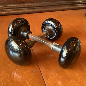 May include: Four antique black glass door knobs with a glossy finish. The knobs are oval-shaped and mounted on aged metal spindles. The door knobs are arranged on a wooden surface, suggesting a vintage or historical theme.