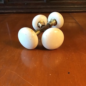 May include: Four antique white porcelain door knobs with brass hardware. The knobs are round and have a slightly aged appearance. The brass hardware connects the knobs, suggesting they are a set. The knobs are resting on a wooden surface.