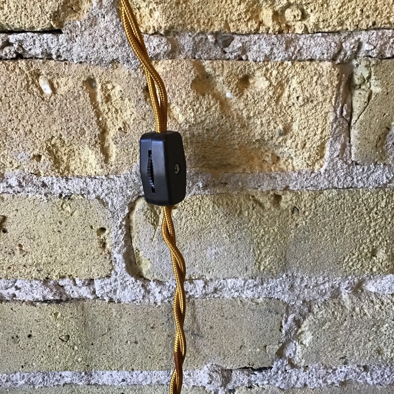 May include: A close-up of a black electrical cord switch on a brick wall. The cord is wrapped in a yellow fabric.