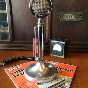 May include: A vintage chrome microphone with a circular mesh head, resting on a round base. Two magazines with orange covers and the text "OST Amateur Radio" are in the foreground. A black meter and a wooden cabinet are in the background.