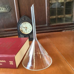 May include: A clear glass funnel with a long, slender spout and a wide, conical body. The funnel is positioned on a brown surface, with a red book and a small clock in the background. The clock has a black frame and a white face.