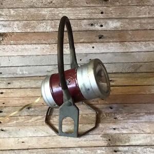 May include: A vintage red and silver spotlight with a metal handle and a tripod stand. The spotlight has a cord and a bulb.