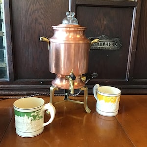 May include: A copper coffee percolator with a glass top and brass accents. Two white mugs with decorative designs sit in front of the percolator. One mug has a green design, and the other has a yellow design.