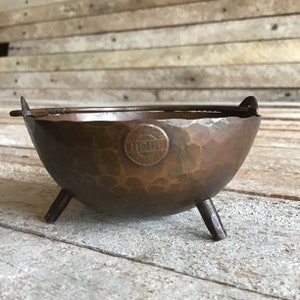 May include: A small, hammered copper bowl with three legs and a handle. The bowl has a circular stamp on the side that reads "HELI".