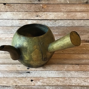 May include: A vintage brass teapot with a rounded body and a long, straight spout. The teapot has a worn, aged patina and is missing its lid.