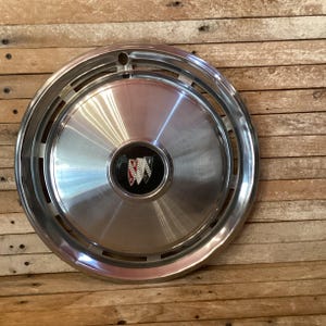 May include: A shiny, silver-colored Buick hubcap with a chrome rim. The center features a black emblem with the Buick logo in red, white, and black. The hubcap is round and has a series of rectangular cutouts around the edge.
