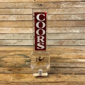 May include: A clear, rectangular Coors beer tap handle with a red and white logo. The handle has a decorative emblem and a metal base. The background is a wooden surface.