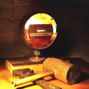 May include: A round, metal gauge with a glass face and a red needle. The gauge is illuminated from behind, and the face reads "HI GAS PRESS". The gauge is sitting on a wooden base, and there is a wooden mallet and a metal tool in the foreground.