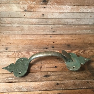 May include: A rustic, antique-style metal handle with a hammered texture and decorative end pieces. The handle is a muted green color and is set against a weathered wooden background.