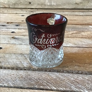 May include: A clear glass cup with a red, ruby-colored rim. The cup has a decorative, raised pattern around the bottom. The rim is etched with the words "Green Edward Inn".