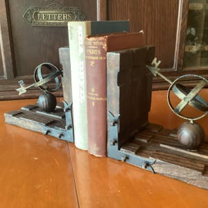 May include: A set of wooden bookends with metal accents, holding several vintage books. Each bookend features a decorative metal armillary sphere and a dark wood base. The books have aged covers and spines, adding to the antique aesthetic.