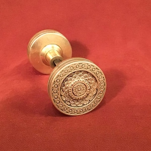 May include: A brass door knob featuring a circular face with an intricate, embossed floral-inspired pattern and a decorative border of small circles. The knob has a smooth, plain cylindrical base and a central metal shaft for installation.