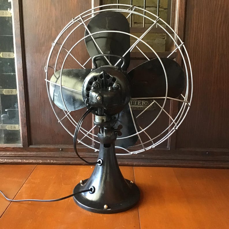 May include: An antique black electric fan with three blades, enclosed in a silver wire cage. The fan has a rounded base and a pivoting head. The brand name "Matters" is visible on the motor housing. The fan is on a wooden surface.