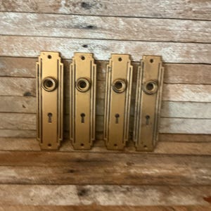 May include: Four rectangular brass door plates arranged in a row. Each plate features a central circular opening for a knob, a keyhole below, and two screw holes at the top and bottom. The plates have a stepped, linear border design.