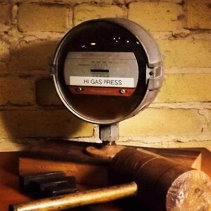 May include: A vintage, round, silver gas pressure gauge with a black face and white lettering. The gauge reads "Hi Gas Press" and has a small, round, red dial with a black needle. The gauge is mounted on a wooden base.