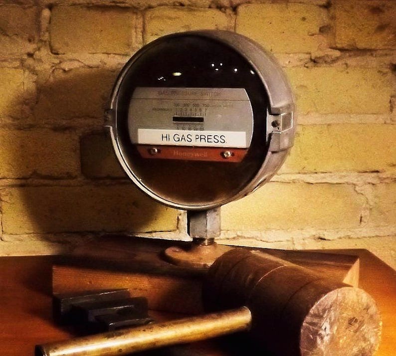 May include: A vintage, round, silver gas pressure gauge with a black face and white lettering. The gauge reads "Hi Gas Press" and has a small, round, red dial with a black needle. The gauge is mounted on a wooden base.