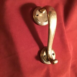 May include: A vintage brass door handle with a curved design. The handle has a rounded grip and is attached to two circular plates with screw holes. The handle is set against a red fabric background.