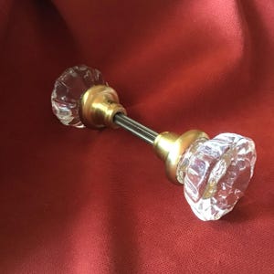 May include: A clear glass door knob with a faceted design and brass hardware. The door knob is connected by a metal rod. The background is a solid red color.