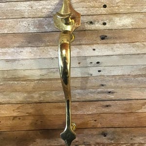 May include: A gold-toned door handle with a decorative design. The handle is curved and has a pointed end.