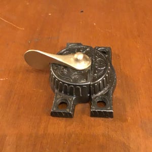 May include: A black metal latch with a brass lever. The latch has an ornate design and is likely for a cabinet or door. The lever is in the open position.