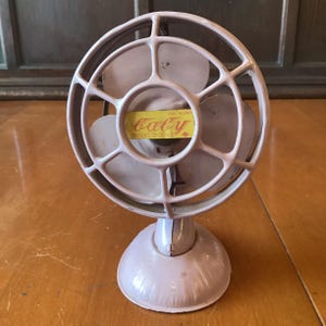 May include: A vintage light pink metal desk fan with a circular protective cage and three blades. A yellow rectangular label on the center of the cage features the word "baby" in cursive script, the text "MADE IN JAPAN", and "PATENT 20092".